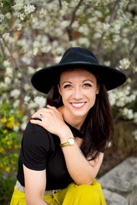 A woman wearing a black felt hat with an oversize brim sits on a low stone wall, legs crossed, elbow propped on her knee. The bush behind her is in bloom, it's sunny, and she's smiling.