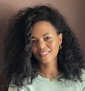 Headshot of women with brown skin, long curly hair in a teal sweater in front of a brown wall.