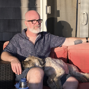 A bald white man with black horn-rimmed glasses, a blue patterned shirt, and a white beard sits on orange patio furniture with a sleeping wheaten terrier lying across his lap.