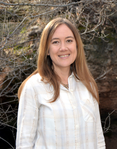 Brown-haired woman dressed in white and gold striped button down shirt, standing in front of tree branches with no leaves
