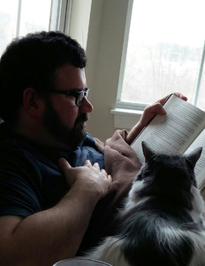 Tim Sheehan sitting in a recliner reading while a cat looks at him