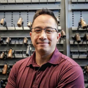 Stephen J. Lu, a middle-aged Asian male wearing a red shirt and glasses, standing in front of a rack of firearms.
