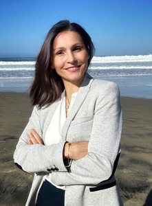 Smiling woman in a gray blazer jacket, standing on a beach with the ocean in the background